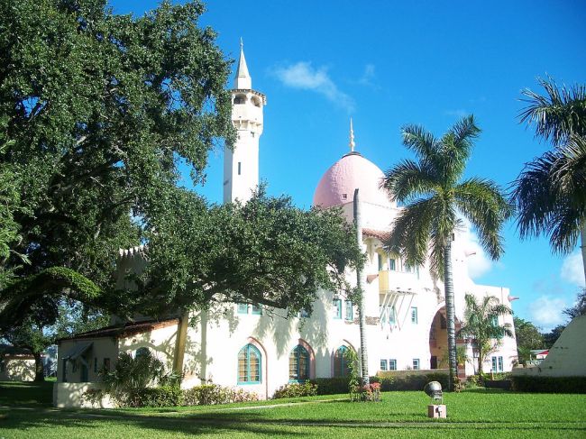 View of a white building with a dome and tower, resembling a mosque, with palms and a live oak in the foreground.